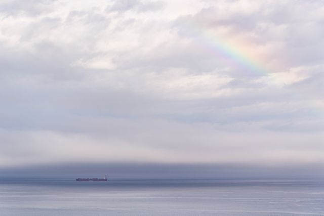 This image showcases a serene ocean view under a partly cloudy sky with a hint of a rainbow. A distant boat floats on the calm waters, creating a peaceful and tranquil atmosphere. Ideal for use in travel magazines, nature websites, and relaxation-themed content. Also great for backgrounds in presentations and blogs about marine life or scenic views.