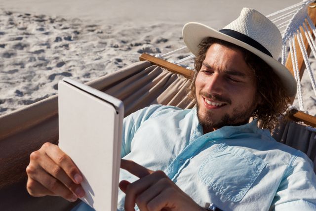 Smiling man relaxing on hammock and using digital tablet on the beach