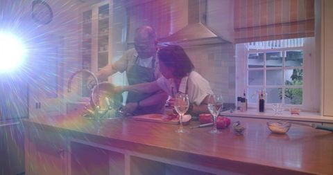 Couple rinsing vegetables at wooden kitchen island while cooking with wine and sunflare