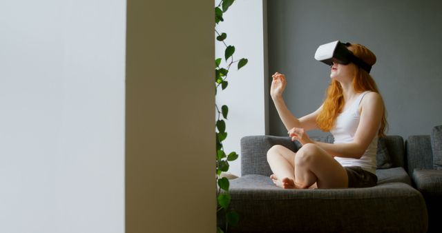Young woman sitting on a sofa using a VR headset in a modern living room. She seems immersed in the virtual experience, moving her hands as if interacting with a digital environment. A green plant on the side adds to the contemporary and comfortable setting. This image is suitable for illustrating technology use in everyday life, advancements in virtual reality, or relaxed home environments.