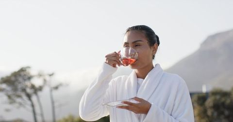 Mid-adult Asian woman wearing white robe sipping pink tea on sunlit balcony, serene morning ritual