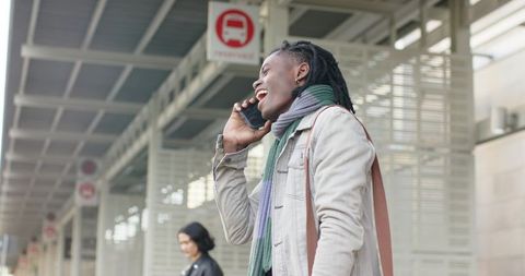 Smiling black woman waiting at bus stop talking on smartphone, urban commuter lifestyle