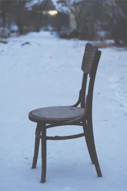 Old wooden chair standing alone in a snowy winter landscape at dusk. Isolated rustic chair in the middle of snow suggests solitude and contemplation. Ideal for use in artistic projects, storytelling, winter-themed designs, or for conveying themes of loneliness and quiet reflection.