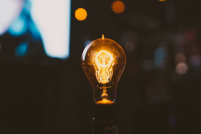 A close-up photograph of an illuminated vintage light bulb with a glowing filament, set against a darkened background that features a beautiful bokeh effect. This image can be used for designs related to creativity, inspiration, energy-efficient lighting, interior design, and industrial decor themes.