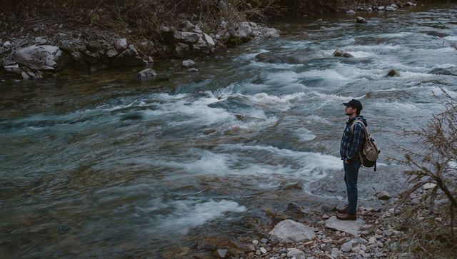 Solo hiker standing on rocky riverbank observing rushing mountain stream under overcast sky