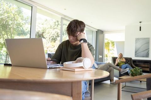 Teenager studying at home with laptop and headphones while parents relaxing in living room
