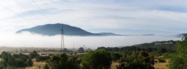 This image depicts a serene morning landscape featuring a distant mountain partially shrouded in fog, set against a field of greenery and clear skies. Ideal for use in nature blogs, travel websites, environmental campaigns, or to evoke a sense of calm and tranquility in advertisements.