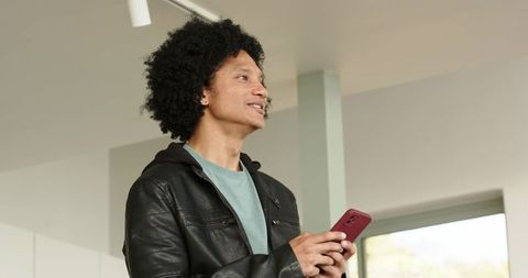 Mid-adult man holding red smartphone in leather jacket, standing in minimalist interior