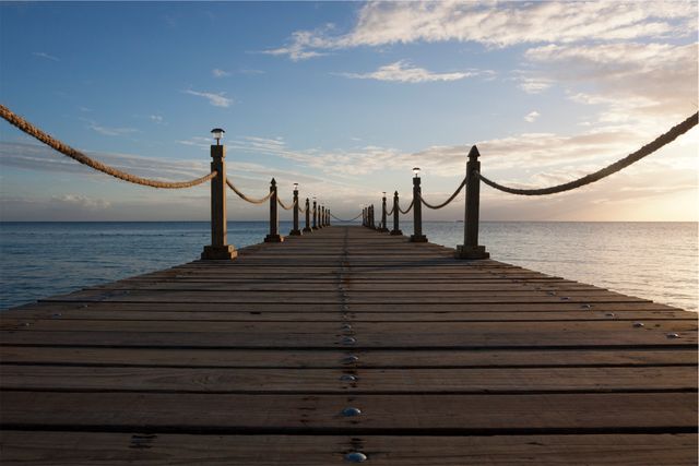 Wooden pier with rope railing extending into smooth sea during sunset. The sky above shows light clouds and warm hues of the setting sun. Ideal for use in travel brochures, vacation advertisements, coastal scenery posters, and for promoting tranquil or serene environments.