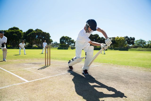 Young man playing cricket on a sunny day with clear sky. He is batting while wearing protective gear, including a helmet. Other players are seen in the background, indicating a team sport. Ideal for use in sports-related content, outdoor activities promotions, and summer recreation advertisements.