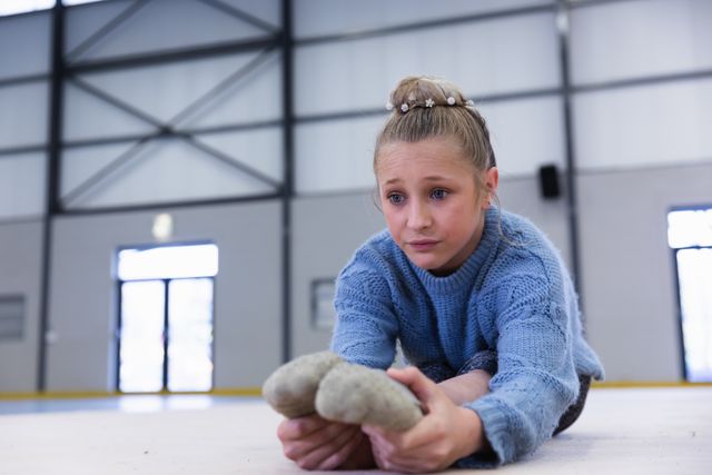 Caucasian female gymnast practicing at the gym, sitting on the floor, stretching her legs and warming up. Gymnast training hard for competition.