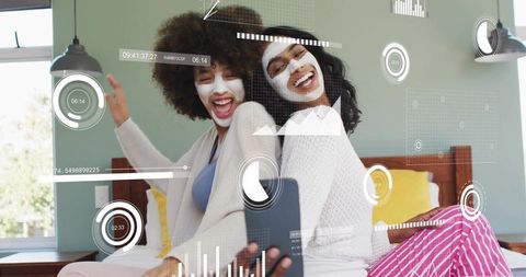 Laughing friends taking skincare selfie with face masks and smartphone in cozy bedroom