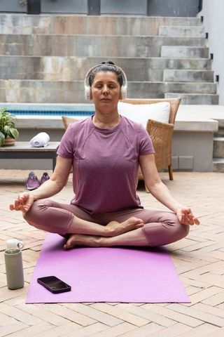 Middle-aged woman meditating with headphones on purple yoga mat by poolside patio