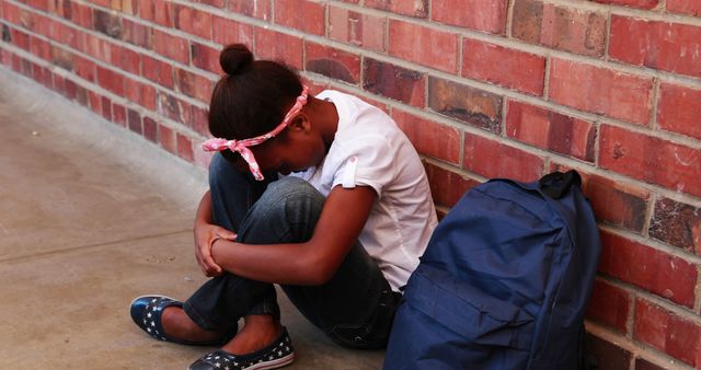 Child sitting alone against brick wall with head down, appearing sad. Backpack placed nearby. Useful for topics on school bullying, stress, depression, loneliness, and mental health in children.