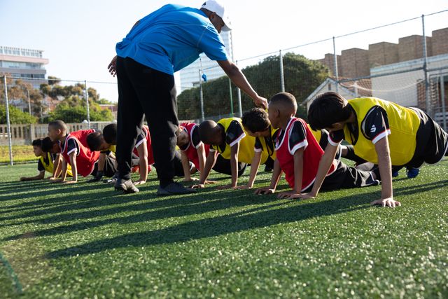 Multi-ethnic children soccer team practicing on a green football pitch on a sunny day, doing press ups, their coach insstructing. Childhood healthy lifestyle competition.