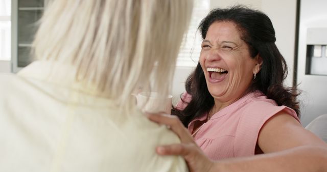 Two women are seen laughing and enjoying a conversation together. One woman is wearing a pink top and is facing the camera, while the other is seen from behind with light-colored hair. This image is perfect for conveying themes of friendship, joy, positivity, and social interaction. Suitable for use in articles about interpersonal relationships, mental well-being, social gatherings, or promotional material for events.