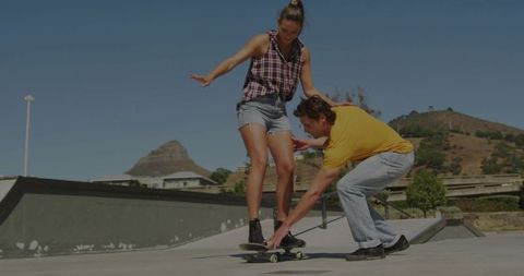 Young woman balancing on skateboard while instructor assisting at sunny urban skatepark