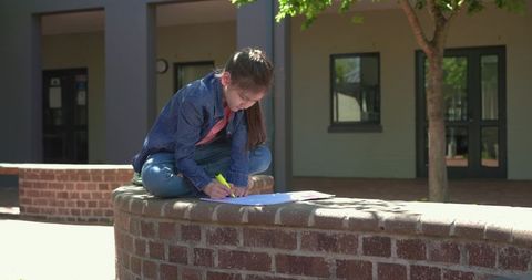 Schoolgirl studying outdoors on brick wall highlighting homework with yellow marker