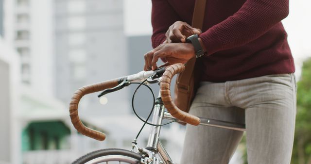 Young man dressed casually in red sweater and jeans viewing his smartwatch while gripping his bicycle's handlebars. This can be used for themes related to contemporary lifestyles, urban commuting, the integration of technology in daily life, and active transportation options for city living.