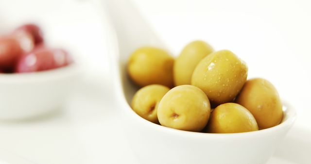 Close-up view of green olives in a white ceramic spoon, highlighting food presentation and freshness. Ideal for use in food blogs, healthy eating articles, or Mediterranean diet promotions.