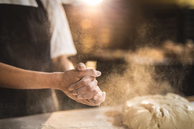 Midsection of female hispanic baker with hands clasped amidst flour and dough in kitchen at bakery. unaltered, blue-collar worker, skilled, food and drink industry concept.