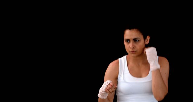 This image features a focused female boxer in a white tank top, performing a fighting stance with hand wraps on a dark background. It is ideal for use in ads, articles, or websites related to fitness, women's empowerment, martial arts training, boxing classes, health and wellness programs, and sports motivation. This visual can also be used for promoting athletic gear or fitness apparel.
