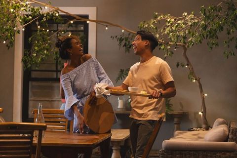 Laughing couple carrying tray and wooden board on cozy patio dining with string lights