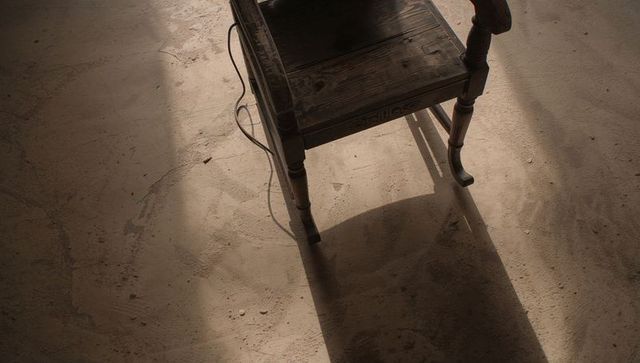Weathered wooden chair casting long shadow on dusty concrete floor with exposed cord