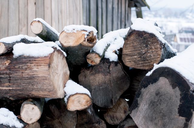 Image shows a stack of firewood logs covered with snow, emphasizing the cold winter season in a rural setting. This is perfect for usage in articles or blogs about winter preparation, rural living, and woodworking in cold climates.