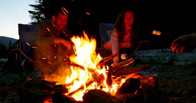 A young Caucasian couple enjoys roasting marshmallows over a blazing campfire at dusk, with copy space. Their relaxed outdoor activity captures the essence of a cozy camping experience.
