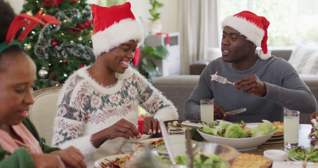 Family gathered around table enjoying Christmas dinner wearing Santa hats. Christmas tree decorated. Smiling and engaging in conversation. Ideal for holiday celebration themes, advertisements, family togetherness content.