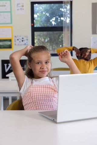 Girl using laptop while african american boy stretching in bright classroom