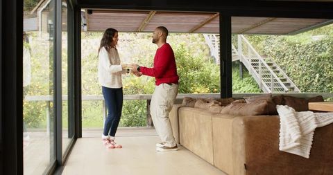 Diverse couple sharing morning coffee in modern living room with glass wall and garden