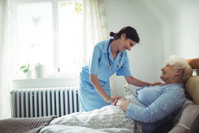 This image shows a nurse interacting with a senior woman who is lying on a bed in a bedroom. The nurse is smiling and appears to be providing care and support, creating a warm and compassionate atmosphere. This image can be used for healthcare, elderly care, home care services, nursing, and medical professional websites or promotional materials.