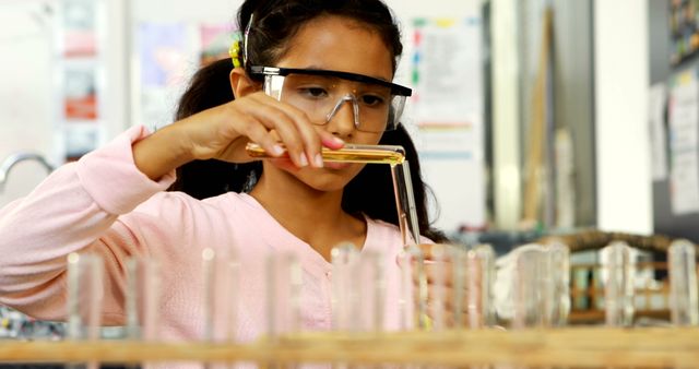 A young girl, of Hispanic or diverse ethnicity, is focused on conducting an experiment in a science class, with copy space. She's wearing safety goggles and carefully pouring a substance into a test tube, showcasing an educational and investigative environment.