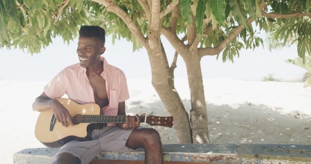 Happy african american man sitting under tree playing guitar and singing on sunny beach, slow motion. Summer, relaxation, hobbies and vacations.