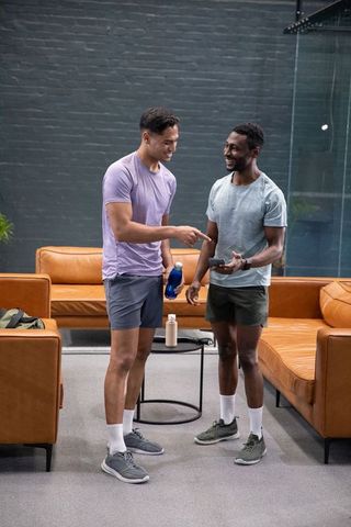 Diverse men chatting and pointing at smartphone in gym lounge holding water bottle