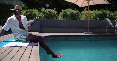 Man relaxing by pool reading paperback with foot in water wearing straw hat