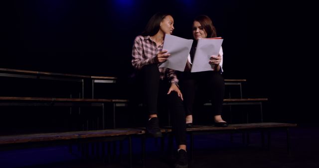 Two women are rehearsing a script while sitting on bleachers on a dark stage. This image can be utilized to represent theatre rehearsals, dramatic arts, teamwork, or script reading. It is useful for articles or content related to performing arts, acting classes, theatre production, or stage performance preparation.