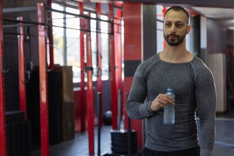 Indian man holding water bottle standing in modern gym near power rack and weight plates