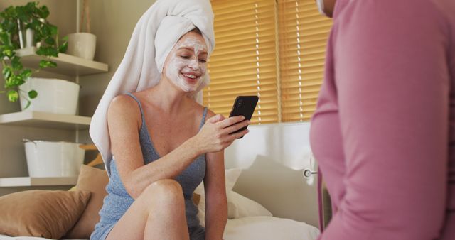 Woman treating herself to a facial mask, sitting on bed, engaging with smartphone. Great for advertising beauty and skincare products, promoting self-care routines, or depicting modern leisure and lifestyle.