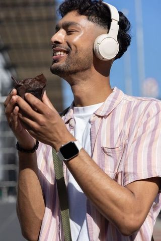 Indian man listening to music with headphones and smartwatch, enjoying sunny urban snack