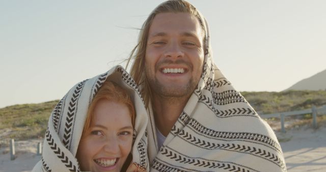 Caucasian couple wrapped in a blanket on the beach. They share a warm embrace, enjoying a romantic moment by the sea.