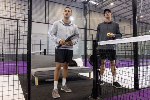 Two men training on indoor padel court holding racket and yellow balls with purple court