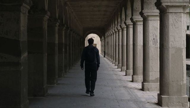 Walking lone traveler down historic stone colonnade with repeating arches and perspective
