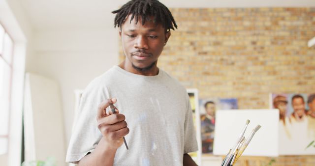 Young male artist in casual gray t-shirt holds paintbrushes in one hand, contemplating his next brush stroke. Background shows an art studio with unfinished canvases and artworks. This image can be used to illustrate the creative process, highlight artistic concentration, or feature a modern painting studio.