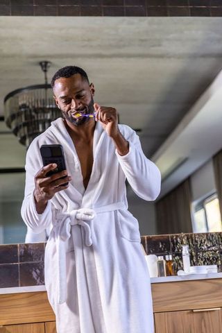 Mature african american man brushing teeth and checking phone in luxury morning bathroom