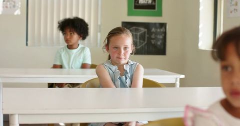 Focused schoolgirl sitting at desk in diverse elementary classroom with classmates