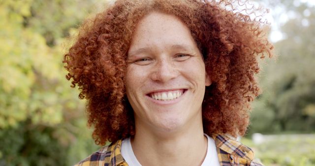 Young man with curly red hair and freckles smiling broadly, wearing plaid shirt. Background features green leaves and blurred nature elements, suggesting a park in autumn. Ideal for use in lifestyle, happiness, and nature-themed publications. Suitable for promoting outdoor activities, fashion for casual styles, or personal wellness concepts.