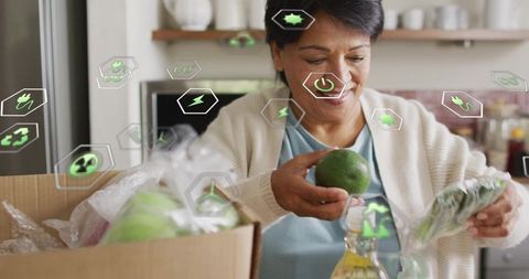 Senior indian woman unpacking groceries holding avocado with eco icons for green delivery
